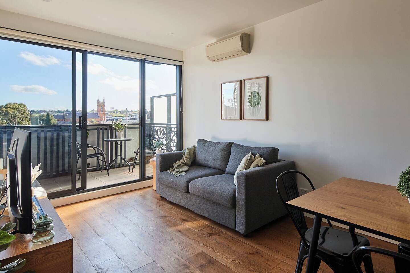 Ein modernes Wohnzimmer mit einem grauen Sofa, einem Holzesstisch und einem Balkon mit Blick auf ein Gebäude.