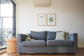 A grey sofa with two cushions, a small wooden side table with a vase and a book, and two framed abstract artworks on the wall. - Urban Drift Cosy Balcony Nook near Waterfront (Geelong)