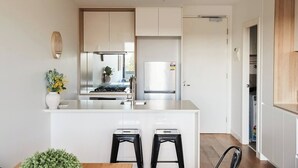A modern kitchen with a central island, two black stools, and a view into the adjacent dining area. - Urban Drift Cosy Balcony Nook near Waterfront (Geelong)
