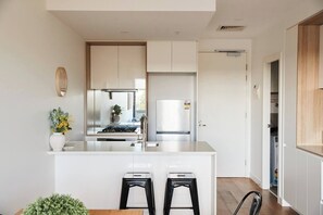 A modern kitchen with a central island, two black stools, and a view into the adjacent dining area. - Urban Drift Cosy Balcony Nook near Waterfront (Geelong)