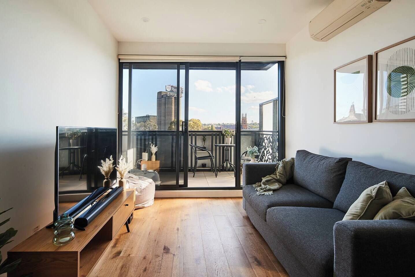 Ein modernes Wohnzimmer mit einem grauen Sofa, einem Holztisch und einer großen Schiebeglasdoor, die auf einen Balkon mit Blick auf die Stadt führt.