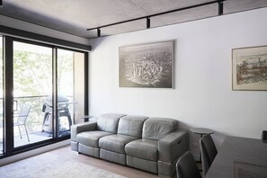 Ein modernes Wohnzimmer mit einem grauen Sofa, einem Esstisch und zwei Stühlen. Ein großes Fenster bietet Blick auf Grünfläche.
