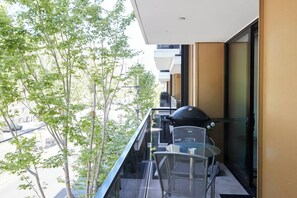 Un balcon avec un barbecue, une table en verre et des chaises, donnant sur une rue bordée d’arbres.