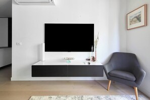 Ein modernes Wohnzimmer mit einem großen Flachbildfernseher an einer weißen Konsole, einem grauen Sessel und einer kleinen Dekorationsvase.