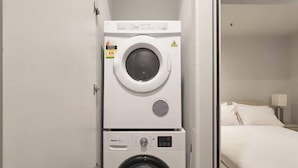 A compact laundry area with a top-loading washer and a front-loading dryer, adjacent to a bed in a hotel room. - Urban Artistry Loft A Haven Above the Bustle (Melbourne)