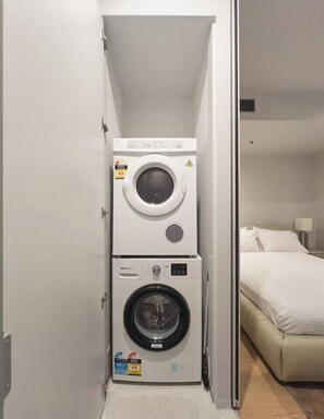 A compact laundry area with a top-loading washer and a front-loading dryer, adjacent to a bed in a hotel room. - Urban Artistry Loft A Haven Above the Bustle (Melbourne)