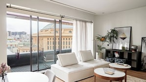 A modern living room with a sofa, a coffee table, and a view of a cityscape through large windows. - Urban Artistry Loft A Haven Above the Bustle (Melbourne)