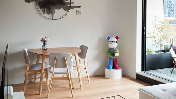 A dining area with a round table and chairs, a wall-mounted abstract art piece, and a window with a view of the outside. - Urban Artistry Loft A Haven Above the Bustle (Melbourne)