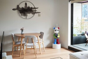 A dining area with a round table and chairs, a wall-mounted abstract art piece, and a window with a view of the outside. - Urban Artistry Loft A Haven Above the Bustle (Melbourne)