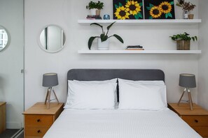 Een keurig opgemaakt slaapkamer met een bed, twee nachtkastjes, een spiegel en wandplanken met boeken en planten.
