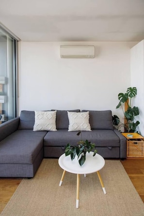 Een moderne woonkamer met een grijze hoekbank, twee witte kussens, een klein rond wit tafeltje met een plant, en een groot raam.