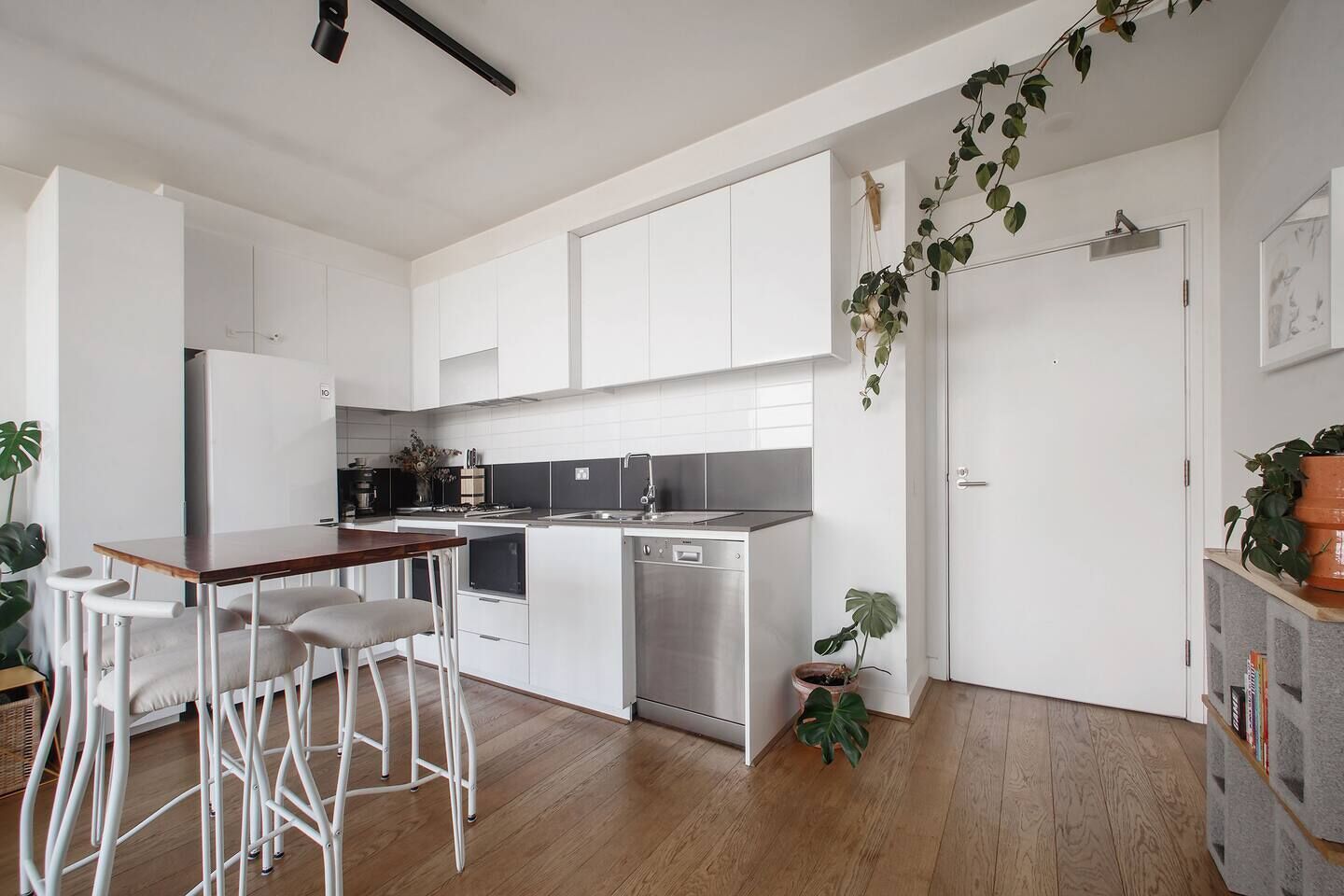 A modern kitchen with white cabinets, a wooden bar counter, and bar stools. There are two large plants, one near the door and one on the counter.