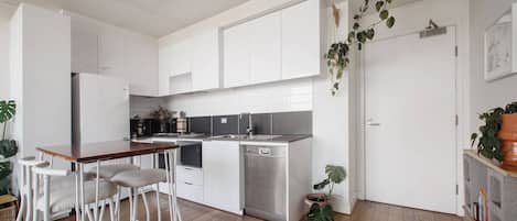 Eine moderne Küche mit weißen Schränken, einer Holzbar und Barhocker. Zwei große Pflanzen stehen dort: eine in der Nähe der Tür und eine auf der Arbeitsplatte.