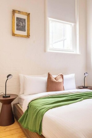 Een slaapkamer met een bed, kussens, een groene plaid, een lijst met een foto aan de muur en een raam.