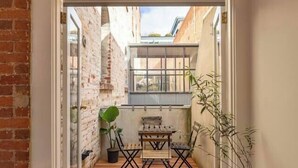 Una terraza de madera con una mesa y sillas, una planta en maceta y vista a un edificio de ladrillo.