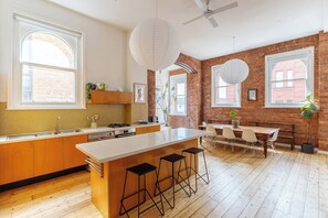 A modern kitchen with a large island, wooden cabinets, and a dining area with a brick wall backdrop. - Foy and Gibson An Industrial Chic Historic Warehouse (Collingwood)