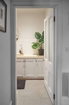 A hallway leading to a bathroom with white cabinets, a sink, and a potted plant. - Nicholson Nook Parkside Stay near Dining and Culture (Fitzroy)