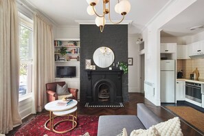 A living room with a fireplace, a television, a bookshelf, and a kitchen area. - Nicholson Nook Parkside Stay near Dining and Culture (Fitzroy)