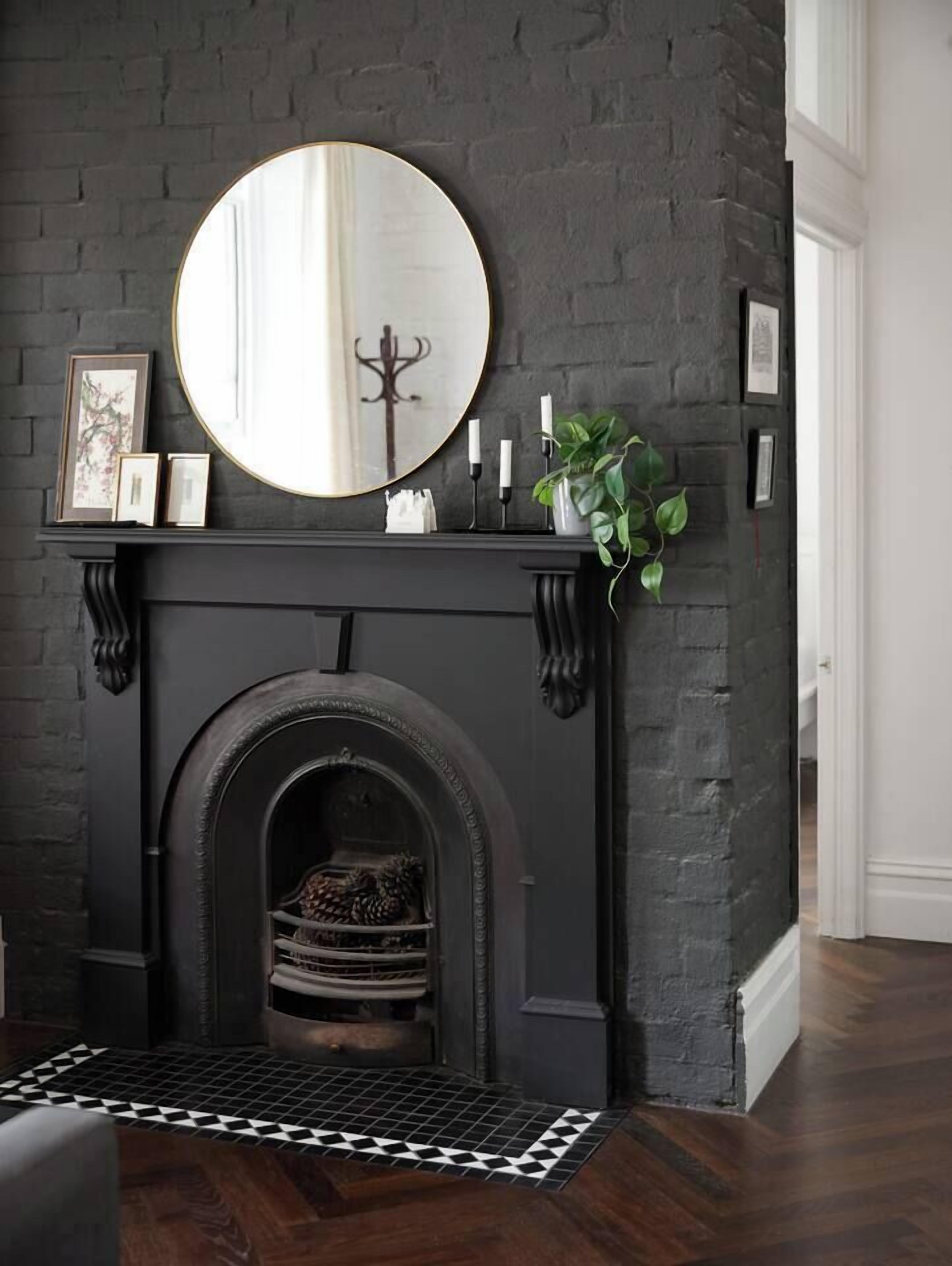 A fireplace with a round mirror above it, a mantel with framed pictures, and a potted plant to the right.