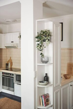 A kitchen with white cabinets, a built-in oven, and a shelf with a plant and decorative items. - Nicholson Nook Parkside Stay near Dining and Culture (Fitzroy)