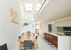 A modern kitchen with a central island, a dining area with a long table, and a staircase leading to an upper level. - Swanston Sands Sophisticated Comfort by the Beach (Geelong)