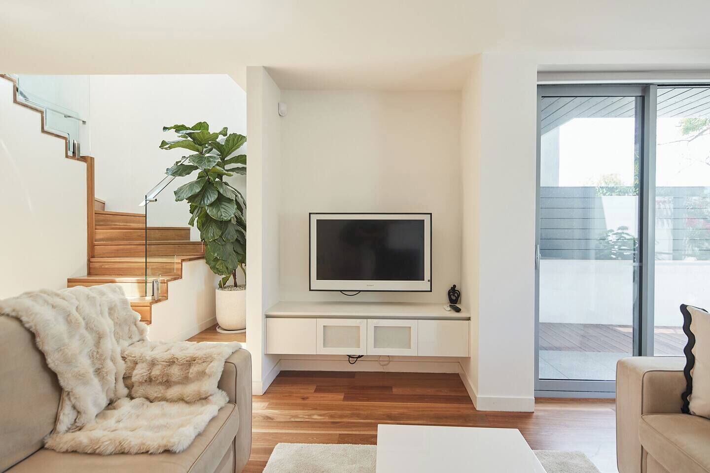 Una sala de estar con un televisor de pantalla plana montado en la pared, una escalera con escalones de madera, una planta grande en maceta y un sofá beige.