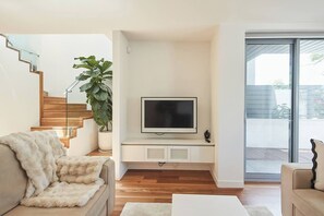 Un saló amb televisió de pantalla plana penjada a la paret, una escala amb graons de fusta, una gran planta en test i un sofà beix.