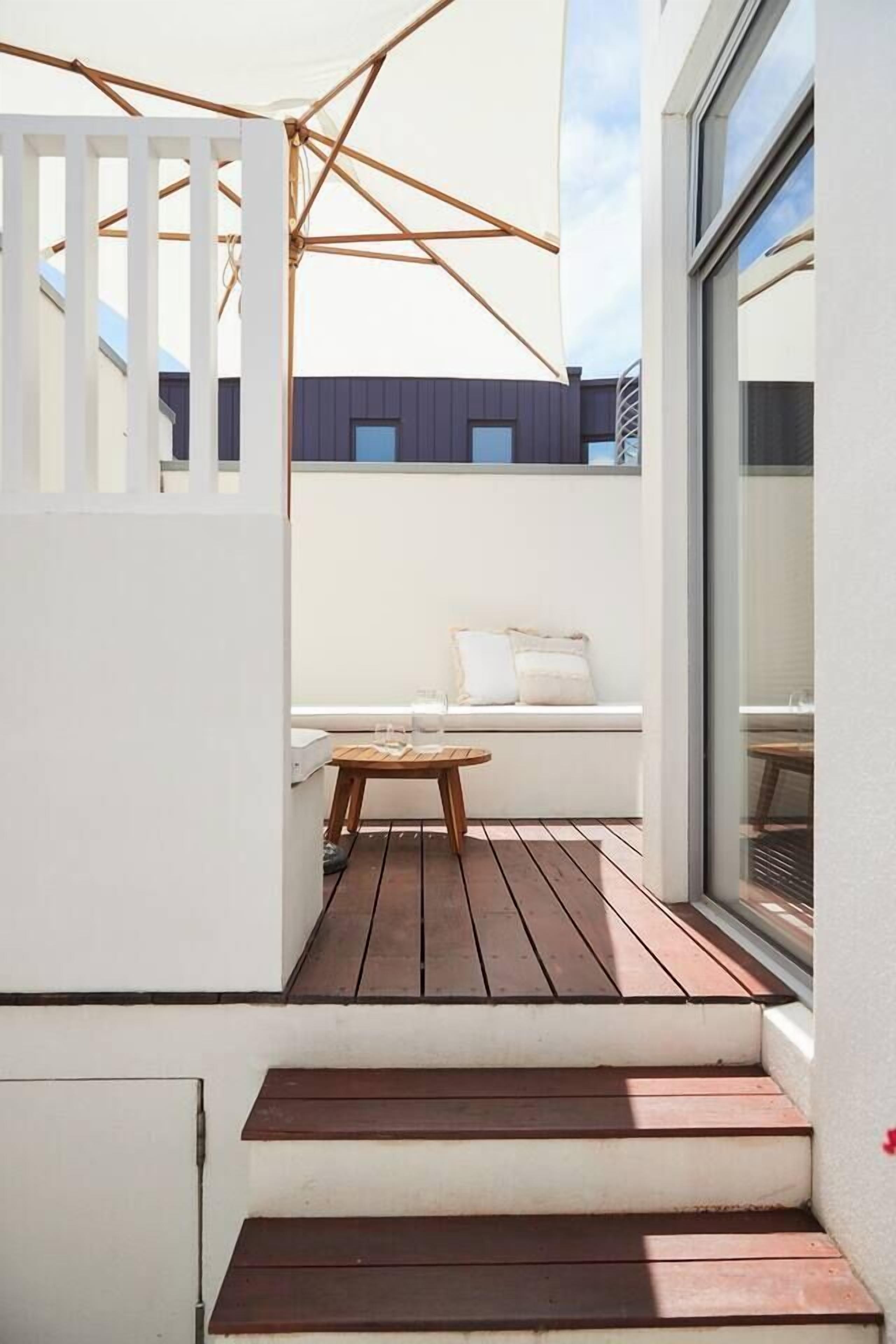 Una terraza de madera con un banco blanco y una mesita pequeña, que conduce a una pared blanca con una ventana.