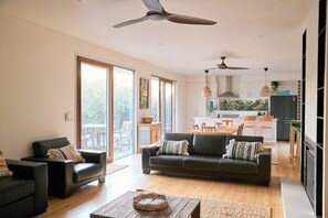 A spacious living room with a dark-colored sofa, a coffee table, and a dining area with a kitchen in the background. - The Garden Escape Slow Living by Beach and Wildlife (Saint Leonards)