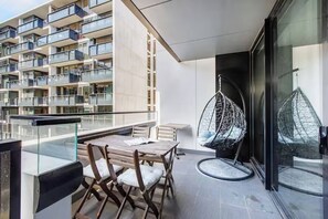 Ein Balkon mit Tisch und Stühlen, einem Hängesessel und einem Glasgeländer mit Blick auf ein modernes Gebäude mit mehreren Balkonen.