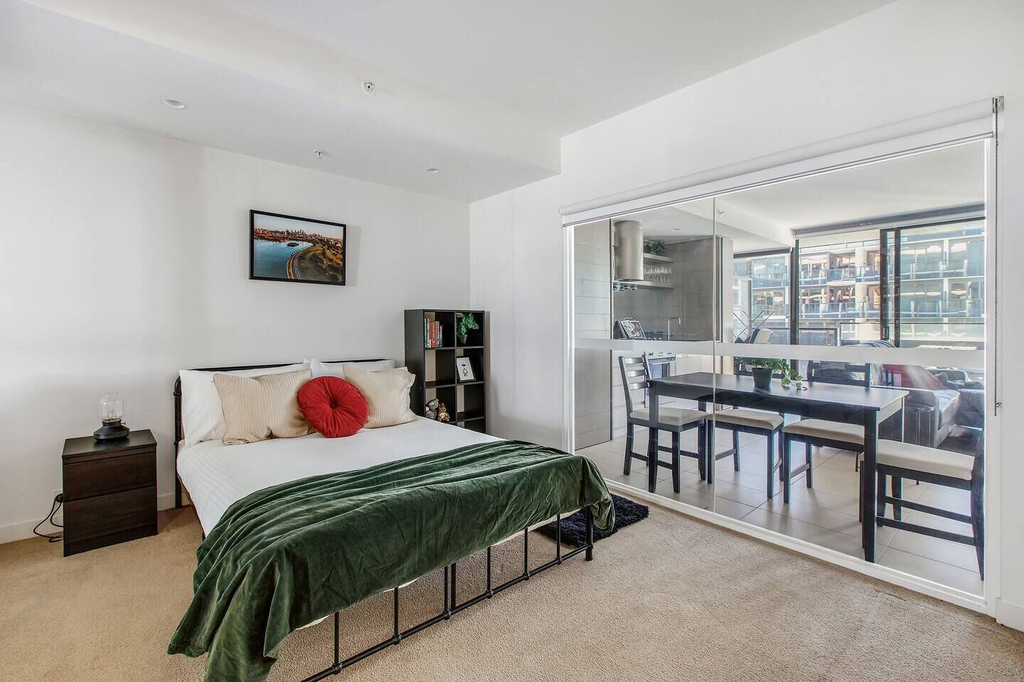 A bedroom with a bed, a nightstand, a bookshelf, and a sliding glass door leading to a balcony with a dining table and chairs.