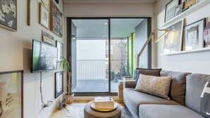 A living room with a sofa, a television, and a bookshelf. There is a sliding glass door leading to an outdoor area. - Urban Escape on Smith Street w Balcony and Parking (Collingwood)