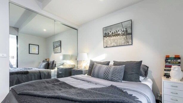 A bedroom with a large bed, bedside tables, a mirror, and a cityscape artwork on the wall. - Urban Escape on Smith Street w Balcony and Parking (Collingwood)