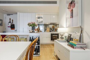 A modern kitchen with white cabinetry, a dining area with wooden chairs, and a small island with a vase of flowers. - Urban Escape on Smith Street w Balcony and Parking (Collingwood)