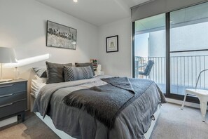Ein Schlafzimmer mit einem großen Bett, einem Nachttisch mit Lampe, einem gerahmten Bild an der Wand und Blick ins Freie durch ein großes Fenster.