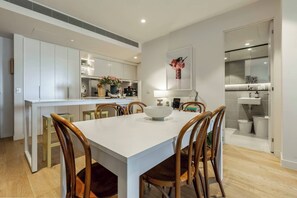 A modern kitchen with white cabinets, a dining table with chairs, and a kitchen island with a bowl. - Urban Escape on Smith Street w Balcony and Parking (Collingwood)
