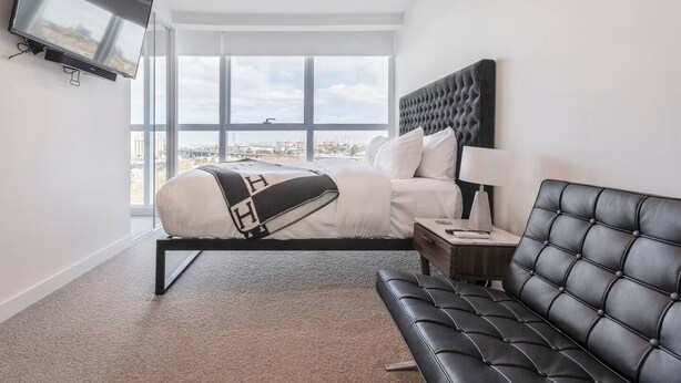 A modern hotel room with a large bed, a black leather sofa, a TV mounted on the wall, and a city view through large windows. - The Fawn Relish in Sub Penthouse Panorama (North Melbourne)