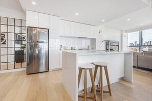 A modern kitchen with white cabinetry, a stainless steel refrigerator, and a bar area with stools. - The Fawn Relish in Sub Penthouse Panorama (North Melbourne)
