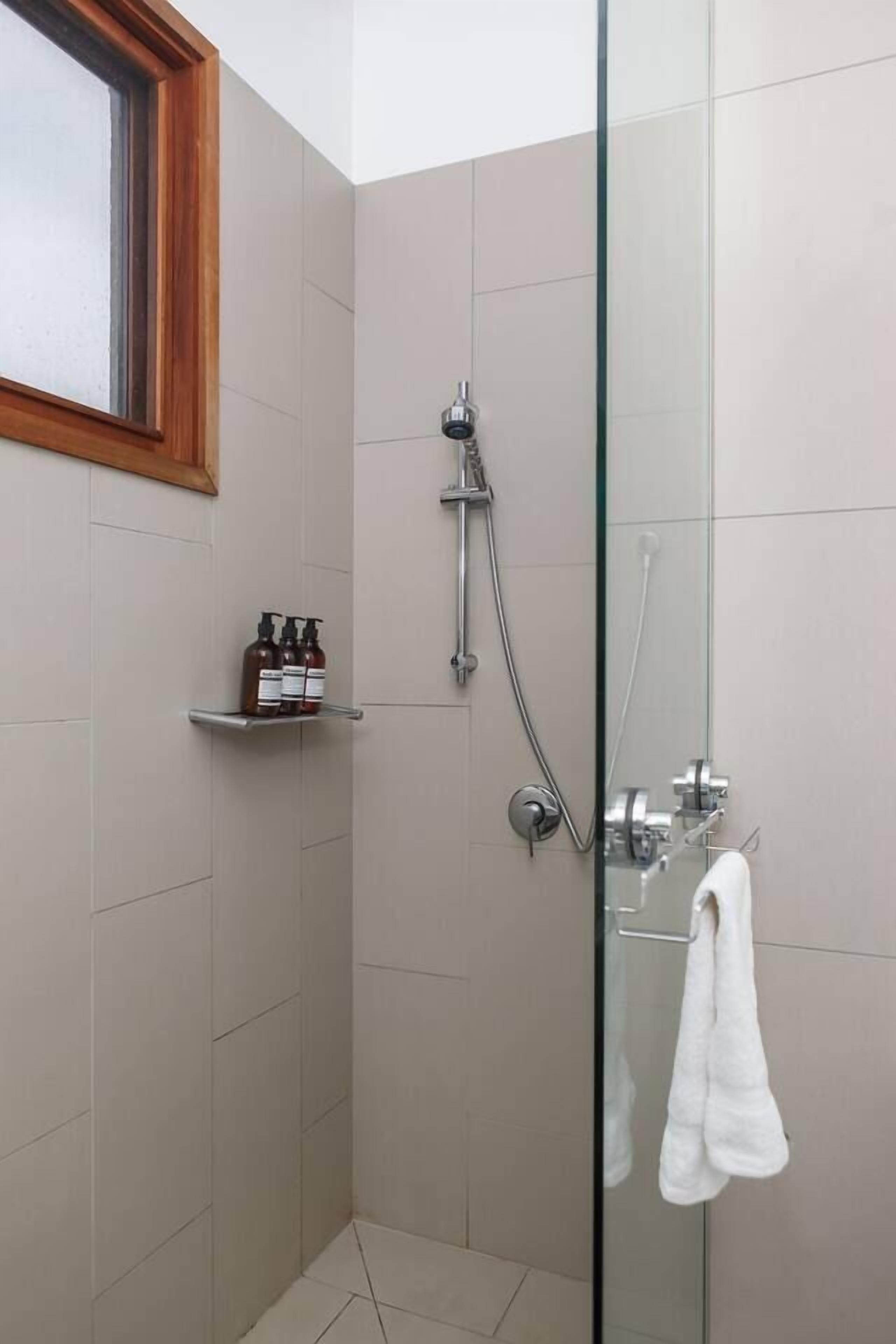 A glass-enclosed shower stall with a towel hanging on the door handle, a shelf with toiletries, and a window above.