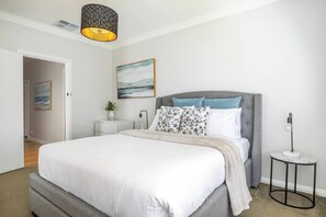 The primary bedroom offers a queen bed with crisp linens, layered pillows, and soothing artwork, creating a tranquil retreat bathed in soft natural light for restful mornings.