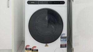 A compact, white washing machine with a front-loading door, a small cabinet to its left, and a power outlet above it. - Northside Rendezvous A Stylish Couples Hideaway (Brunswick East)