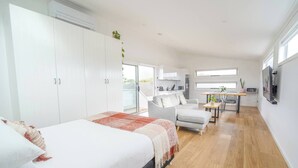 A modern, well-lit bedroom with a large bed, a seating area with a sofa and chairs, and a kitchen visible through an open door.
