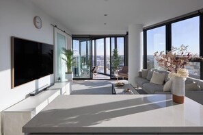 A modern living room with a large flat-screen TV, a sofa, a coffee table, and a view of the city through large windows. - Panoramic Elegance Stylish off Chapel St with Pool (South Yarra)