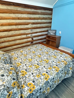 A room with a wooden headboard, a bed with a floral bedspread, a small wooden shelf, and a wall with a blue accent.