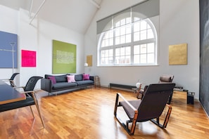 A modern room with a wooden floor, a sofa, a desk, and a large window. - Hoffman Square - Two Bedroom Apartment, Sleeps 4 (London)