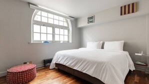 A bedroom with a large bed, a window, a small table, and a bedside table with books. - Hoffman Square - Two Bedroom Apartment, Sleeps 4 (London)