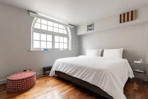 A bedroom with a large bed, a window, a small table, and a bedside table with books. - Hoffman Square - Two Bedroom Apartment, Sleeps 4 (London)