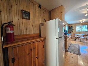 A kitchen with wooden walls, a white refrigerator, a fire extinguisher, and a dining area with chairs and a table.