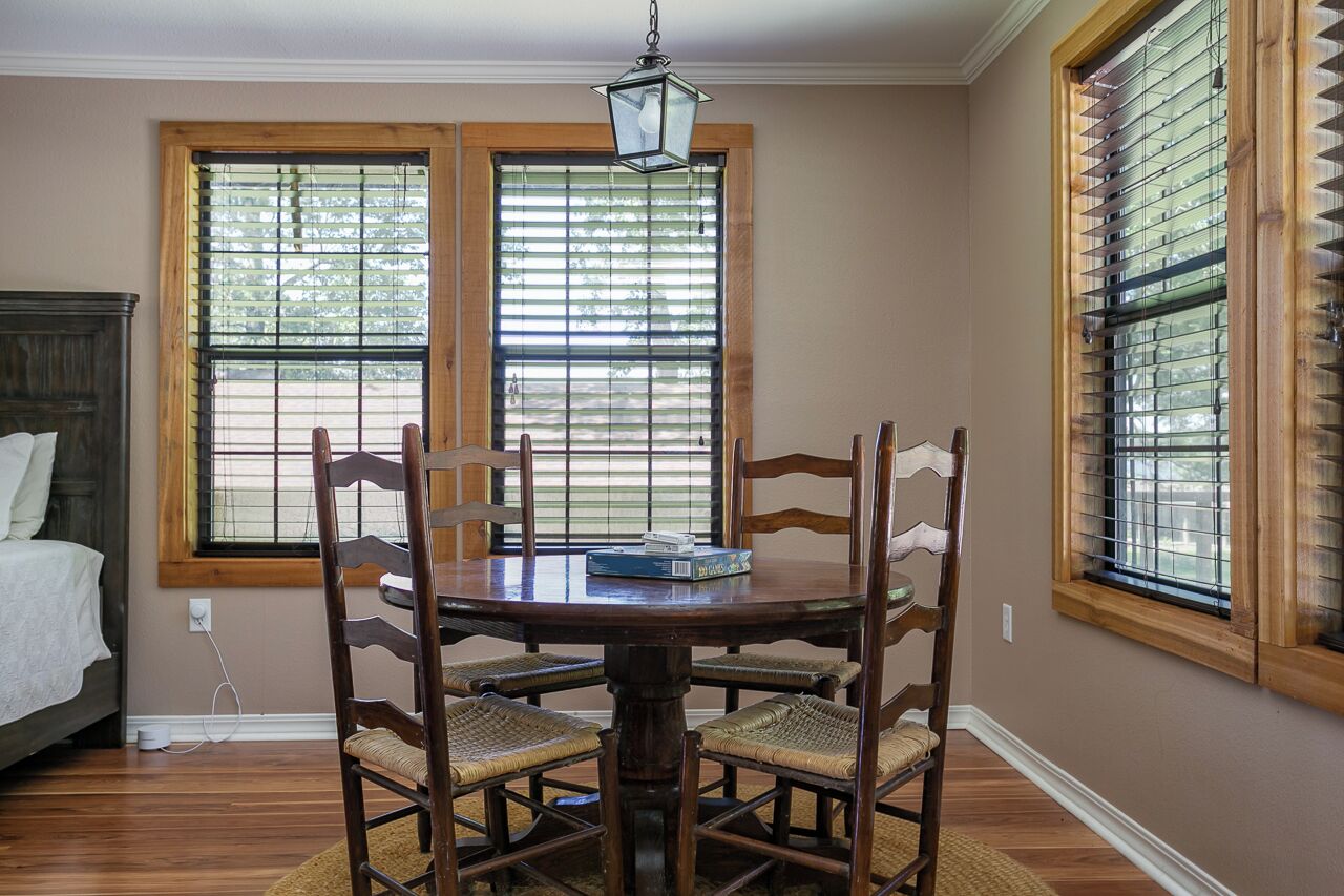 A dining area with a wooden table and chairs, a chandelier, and large windows with blinds.
