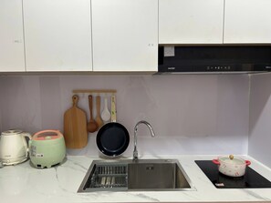 Una cocina con un lavamanos de acero inoxidable, una tetera, una cocedora de arroz y una olla sobre una placa de inducción.
